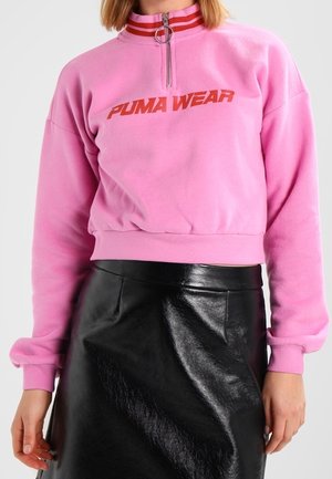 Woman wearing a pink Puma Wear sweatshirt with a red-striped zipper collar and a black shiny leather skirt.