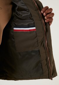 Brown quilted jacket interior with a side zipper, featuring an inner pocket with a red, white, and blue striped accent. Smooth texture.