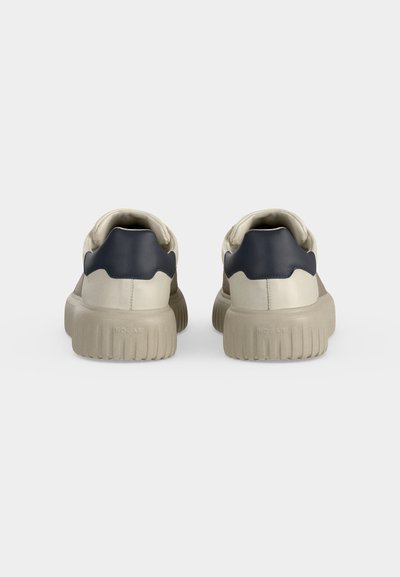 Paire de baskets beiges avec semelles épaisses et panneaux noirs au talon vues de dos sur un fond clair uni.