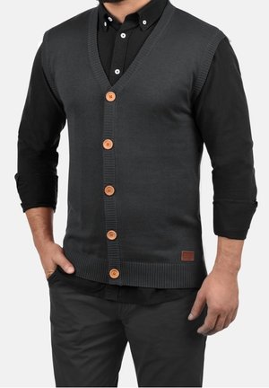 Mężczyzna ubrany w ciemnoszary, zapinany na guziki bezrękawnik, na czarnej koszuli z długim rękawem i czarnych spodniach, z częściowo wsadzonymi rękami do kieszeni.