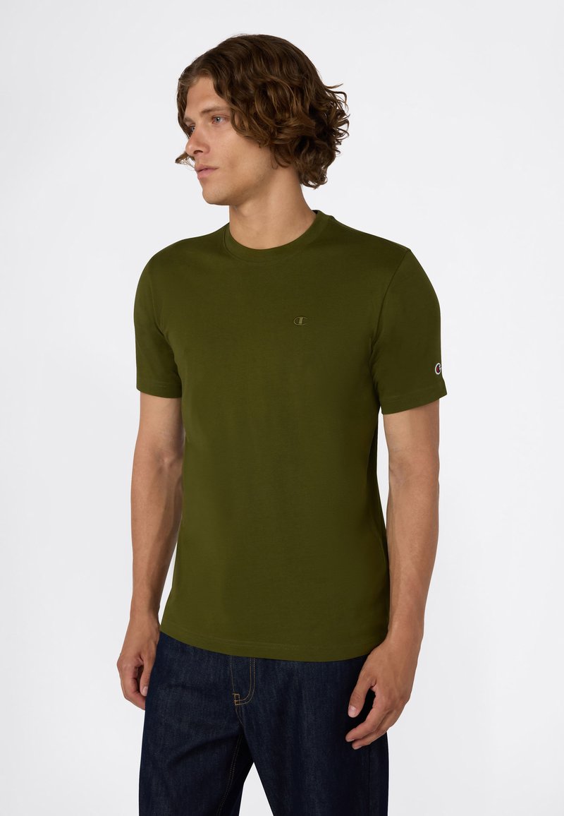 Jonge man met krullend haar, gekleed in een olijfgroene T-shirt met korte mouwen en donkerblauwe jeans, die naar links kijkt tegen een effen achtergrond.