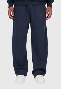 Person trägt locker sitzende, marineblaue Sweatpants und weiße Sneaker, steht mit entspannten Händen an den Seiten vor einem einfarbigen Hintergrund.