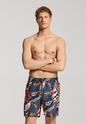 Shirtless man with arms crossed wearing colorful patterned swim shorts and a shell bracelet, standing against a plain background.