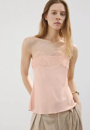 Donna che indossa un top color rosa chiaro con dettagli in pizzo e pantaloni beige plissettati, in piedi davanti a uno sfondo chiaro e semplice.