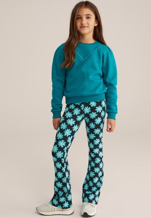 Felpa turchese con un motivo a rombi di borchie, abbinata a pantaloni neri a zampa con grandi fiori blu. Sneakers bianche.