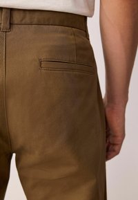 Bruine broek met een gladde textuur, rechte snit en één achterzak. De stof lijkt duurzaam en lichtgewicht.