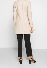 Cappotto beige a maniche lunghe con taglio dritto, spacchi laterali e tessuto leggermente strutturato, abbinato a pantaloni neri a sigaretta e tacchi neri chunky.