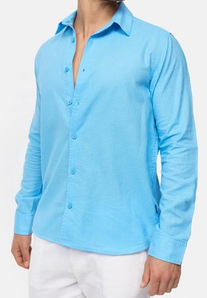 Mann trägt ein hellblaues, langärmeliges Hemd mit offenem Kragen und weiße Hosen, steht vor einem schlichten Hintergrund.