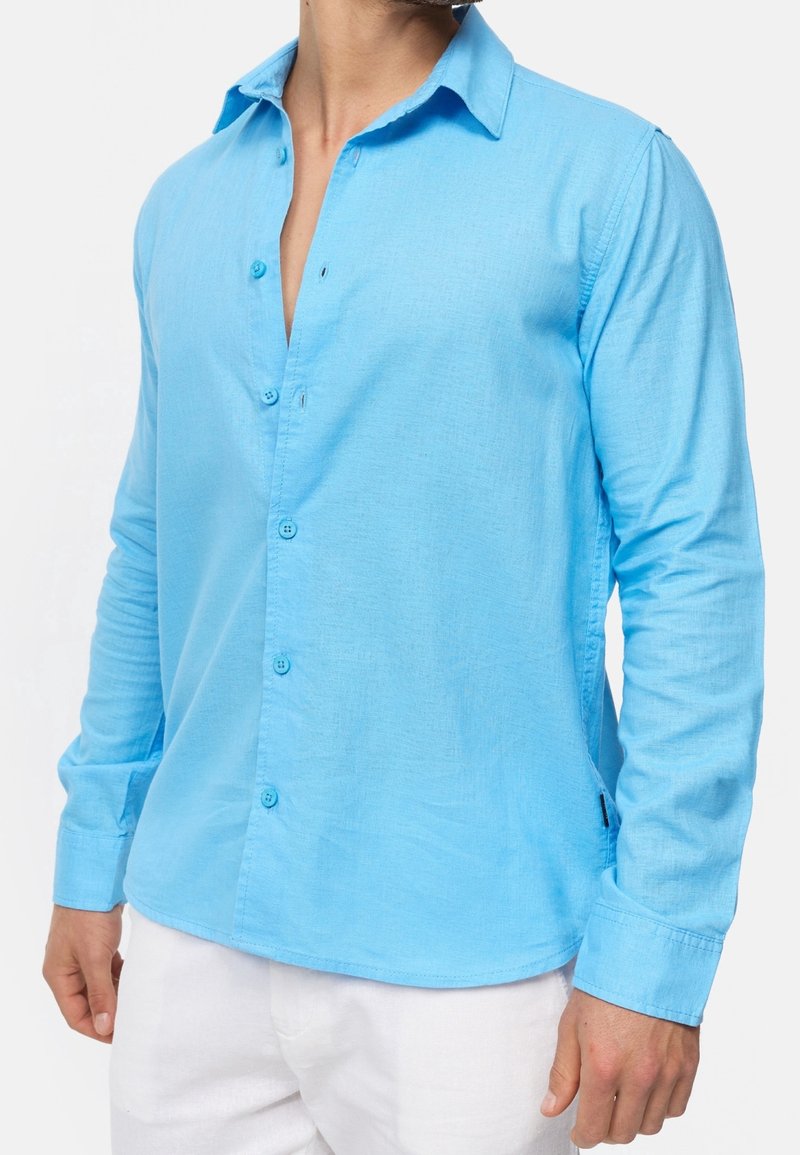 Homme portant une chemise à manches longues bleu clair avec col ouvert et un pantalon blanc, debout devant un fond uni.