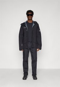 Zwarte waterdichte jas met grijze accenten, voorzien van een capuchon, ritszakken en verstelbare manchetten. Gecombineerd met zwarte broek en schoenen.