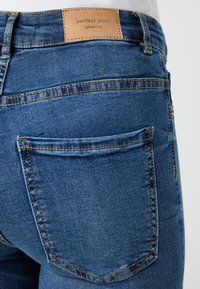 Dark blue denim jeans with a textured finish, featuring a leather label reading "perfect jeans," and visible stitching on pockets.