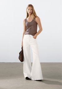 Lichtbruine geribde tanktop gecombineerd met hoge witte flare broek met een vetersluiting aan de voorkant. Model houdt een bruine gevlochten handtas vast.