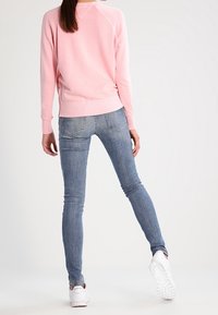 Hellrosa Pullover mit geripptem Saum, kombiniert mit eng anliegenden blauen Jeans. Das Outfit wird durch weiße Sportschuhe abgerundet.