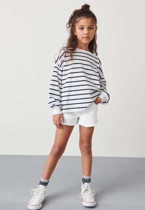 Ung pige står med den ene hånd i lommen, iført en hvid langærmet stribet trøje, hvide shorts, stribede sokker og hvide sneakers.