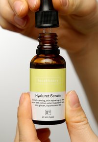 Bouteille en verre brun avec un capuchon compte-gouttes noir. Étiquette blanche et vert clair indiquant le nom du produit et les ingrédients. Doigts du titulaire visibles.