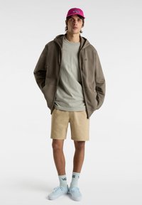 Brauner Reißverschluss-Hoodie, hellgrünes T-Shirt, beigen Shorts, pinke Kappe, blaue Schuhe und hellblaue Socken mit einem Logo. Einfaches, lässiges Outfit.
