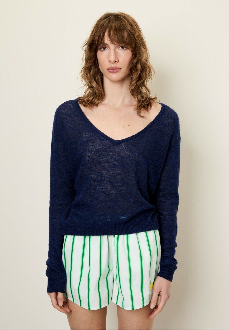 Jūrinio mėlynumo megztinis su V formos kaklu ir ilgomis rankovėmis, derinamas su baltais šortais, kurie turi žalius vertikalius dryžius ir geltoną akcentą.