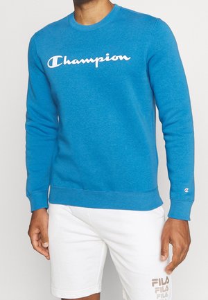 Mann trägt ein leuchtend blaues Champion-Sweatshirt mit weißem Logo vorne und weiße FILA-Shorts mit Logo am linken Bein, steht vor einem einfarbigen Hintergrund.