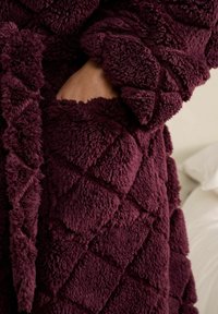 En persons hånd inde i lommen på en dyb lilla tekstureret fleecejakke med et diamantmønster, mod en lys baggrund.