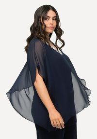Marineblaue transparente Bluse mit kurzen Ärmeln, die einen V-Ausschnitt mit dekorativen Akzenten und einem fließenden Design aufweist.