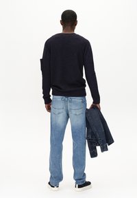 Mornarska pulover z okroglim izrezom, v kombinaciji s svetlim jeansom. Oseba drži temno denim jakno, obuta ima črno-bele superge.