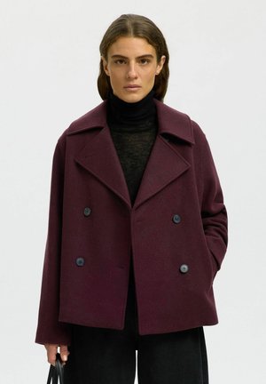 Femme portant un manteau croisé violet foncé sur un col roulé noir, tenant un sac à main noir, une main dans la poche.