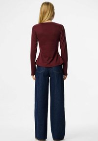 Sieviete ar gariem blondiem matiem, tērpusies pielāgotā bordo garās piedurknes topā un platās tumši zilās biksēs, stāv pretējā virzienā uz balta fona.