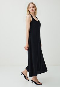 Czarna, bezrękawnikowa sukienka maxi z okrągłym dekoltem, gładką teksturą i zwiewnym fasonem. Noszona z czarnymi klapkami na obcasie.