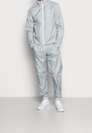 Personne portant un survêtement Nike gris clair avec des passepoils blancs et des baskets blanches, debout devant un fond clair uni.