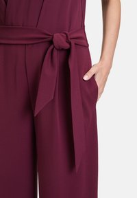 Combinaison bordeaux avec une ceinture nouée à la taille, présentant un tissu lisse et texturé, ainsi que des poches latérales.