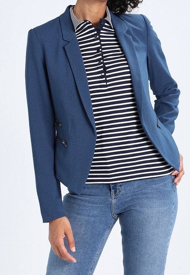 Femme portant un blazer bleu sur un polo rayé noir et blanc avec un jean bleu, main posée sur la hanche.