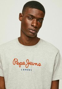 Pilki trumpi marškinėliai su oranžine kursyve užrašyta "Pepe Jeans" ir "LONDON" mėlyna spalva. Lygus medvilnės audinys su laisvu kirpimu.