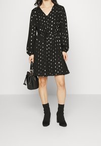 Černé šaty s stříbrnými puntíky, véčkovým výstřihem, dlouhými rukávy a zavazováním v pase. V kombinaci s černými botami na podpatku a malou černou taškou.