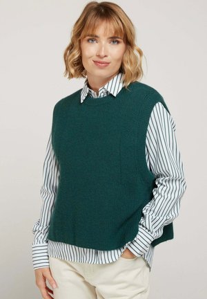 Pull sans manches en tricot teal avec un col ras du cou, finition texturée, porté par-dessus une chemise blanche et verte à rayures. Tissu léger, coupe décontractée.