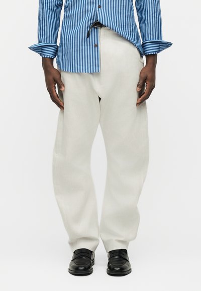 Άνδρας που φοράει άνετο λευκό παντελόνι, μαύρα επίσημα παπούτσια και μπλε πουκάμισο με κάθετες ρίγες και κουμπιά, με τα μανίκια σηκωμένα.