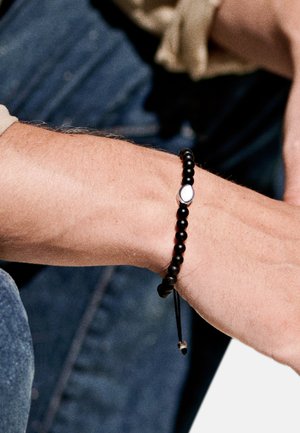 Svart pärlarmband med en metallisk oval detalj och slät textur. Justerbar snodd för passform. Hand med armbandet synligt.