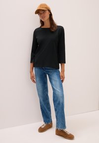 Frau mit braunem Hut, schwarzem 3/4-Ärmel-Oberteil, blauen Jeans und braunen Turnschuhen, die vor einer schlichten hellen Wand steht.