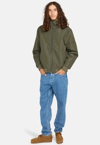 Olijfgroene zip-up jas met een hoge kraag, gecombineerd met lichtblauwe baggy jeans en bruine suède schoenen. Heeft een minimalistisch design.
