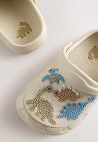 Sabots béiges pour enfants avec des motifs de dinosaures colorés en relief sur le devant et de petites illustrations de dinosaures à l'intérieur des talons.
