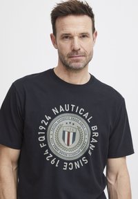 Sort bomulds T-shirt med et cirkulært logo med laurbærblade og teksten: "FQ1924 NAUTICAL BRAND SINCE 1924" i grå og hvid.