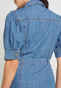 Denimmekko puff-hihnoilla, vaaleansininen väri, vyötäröltä kiristetty ja kauluksellinen. Sisältää napitettu hihansuun yksityiskohtia ja näkyvää ompelutyötä.