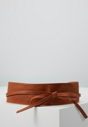 Large ceinture en cuir marron avec une fine lanière nouée en nœud à l'avant, présentée sur une surface blanche avec un fond uni.