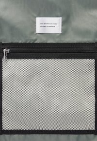 Gray recycled fabric pouch with a black zipper. Features a mesh front pocket, showcasing a textured pattern and a tag stating its materials and design origin.