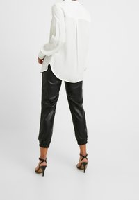 Persona che indossa una blusa bianca larga, pantaloni jogger in pelle nera con polsini elasticizzati e sandali neri con tacco alto, in piedi su un pavimento bianco.