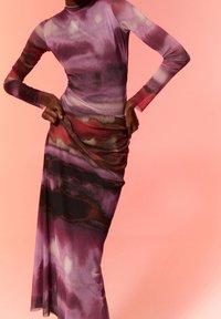 Personne portant une robe longue à manches longues avec des motifs abstraits violets, roses et rouges, tenant la jupe à la hanche sur un fond rose.