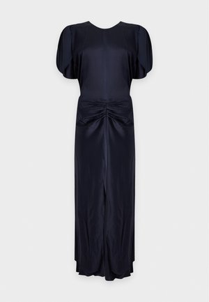 Femme aux longs cheveux foncés portant une robe en satin noire à manches, avec des fentes aux épaules et des fronces à la taille, associée à des talons noirs ouverts.