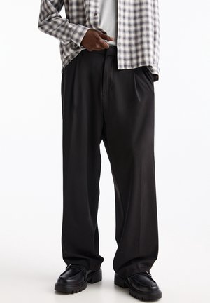 Person, die schwarze, weit geschnittene Hosen, schwarze klobige Loafer, ein weißes Hemd und ein graues kariertes Hemd mit Knöpfen trägt.