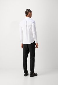Camisa de manga longa branca com colarinho abotoado, tecido suave e bainha curva, combinada com calças pretas e sapatos escuros.
