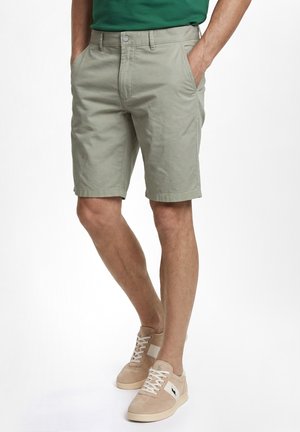 Mann trägt beige knielange Shorts, grünes Hemd und hellbeige Sneakers mit weißen Schnürsenkeln, steht mit den Händen in den Taschen.