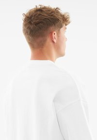 Bílý bavlněný sweatshirt s volným střihem, oblečeným kolem krku a spadlými rameny, s hladkým, jednobarevným materiálem bez vzoru.